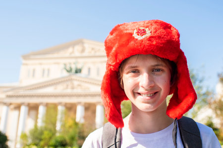 Portrait of a teenage boy in a red ushanka hat with a USSR crest, a child in a white T-shirt with a backpack stands in front of the Bolshoi Theater - a landmark of Moscow.の写真素材