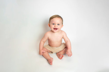 Smiling baby boy in beige shorts sitting on a white background. The child is 9 months old.