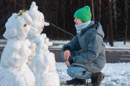Teenage boy in gray jacket and green hat sitting facing small snowmen in winter, side view.の写真素材