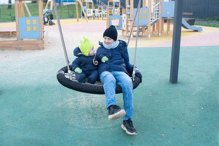 A teenager and his younger brother ride on a nest swing at a playground in late autumn.