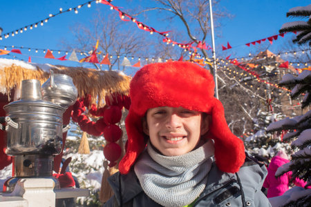 happy teenage boy in red ushanka hat with samovar at winter festival in Moscow