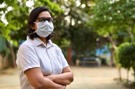 Side angle portrait of a young medical healthcare female worker with hands crossed / folded, wearing surgical mask to protect herself from Corona Virus (COVID-19) pandemic against blur backgroundの写真素材