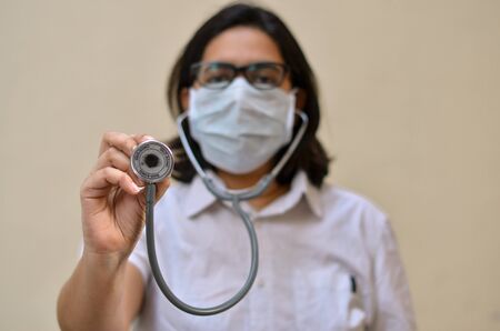 Portrait of young medical healthcare female worker showing / holding out her stethoscope & wearing surgical face mask to protect from Corona Virus (COVID-19) pandemic against yellow backgroundの写真素材