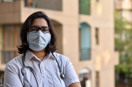 Portrait of young medical healthcare female worker with hands crossed / folded, stethoscope over her shoulders wearing surgical mask to protect herself from Corona Virus (COVID-19) pandemic diseaseの写真素材