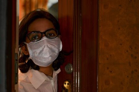 Concerned & worried lady under lock down home quarantine wearing protective breathing surgical face mask against flu and novel Corona virus (Covid-19) pandemic & greeting visitor through door / windowの写真素材