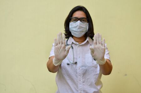 Portrait of young medical healthcare female worker showing her hands out & wearing surgical face mask to protect from Corona Virus (COVID-19) pandemic against yellow background.Concept Ready to fightの写真素材