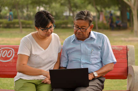 Young Indian woman manager in western formals / shirt helping old Indian man on a laptop promoting digital literacy for elderly in a park in New Delhi, India.Concept: father daughter/ Digital literacyの写真素材
