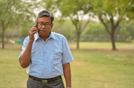 Senior businessman or working professional Indian man wearing a half sleeve shirt and trousers standing in a park speaking on his smart phone in Delhi. Concept digital literacy with senior citizensの写真素材
