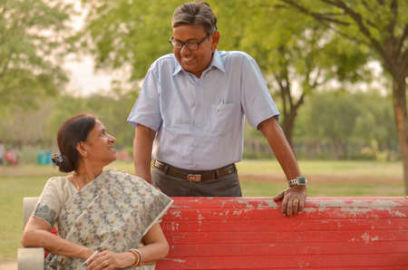 Happy looking retired senior Indian man and woman couple, sitting on a red bench, smiling and looking at each other in a park outdoor in New Delhi, Indiaの写真素材