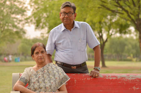 Happy looking retired senior Indian man and woman wearing saree couple smiling sitting on a red park bench in an outdoor setting in New Delhi, Indiaの写真素材