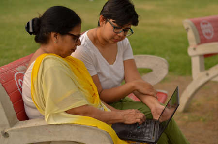 Side view of young Indian girl helping an old Indian woman on a laptop sitting on a red bench in a...