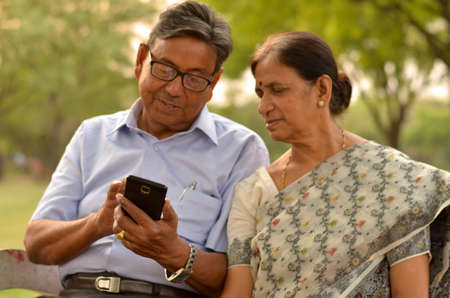 Portrait of senior couple sitting in park bench and looking at their smart phone and laughing in New Delhi, India with focus on the handsの写真素材