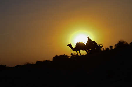 Silhouette of a camel cart and camel carrying tourists in Sam sand dunes, Jaisalmer. Located in the...