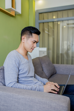 Asian man at home on sofa using a laptop.の写真素材