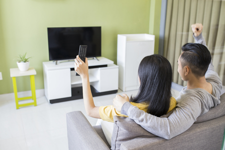 Happy asian couple watching a movie on tv sitting on a couch at homeの写真素材