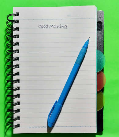 Note Book with Pen of Sky Color on a Green background. Saying Good morning.