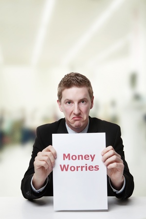man in a suit sat at a desk look at a  piece of paper with the words money worries printed on itの写真素材