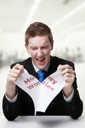 man in a suit sat at a desk ripping a  piece of paper up with the words money worries printed on it and looking happy doing so.の写真素材