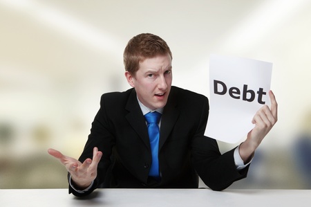 man in a suit sat at a desk look at a  piece of paper with the word debt printed on itの写真素材