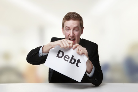 man in a suit sat at a desk ripping a  piece of paper up with the word debt printed on it and...