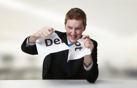 man in a suit sat at a desk ripping a  piece of paper up with the word debt printed on it and looking happy doing so.の写真素材