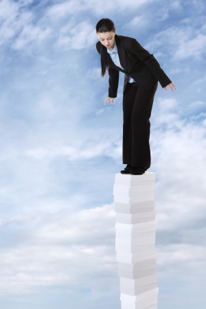 business woman standing on a very large stack of paper work, going high up into a sky  and she looking down at the earth below herの写真素材