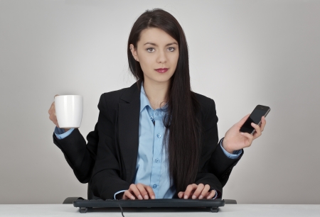 woman sitting at her desk with four arms two hands typing onto a keyboard and one holding a mobile phone and the other a cup of coffee or teaの写真素材