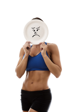 woman wearing sport bra and shorts holding a paper plate with a sad face draw on the plate over her faceの写真素材