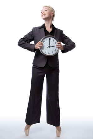 Studio shot of a pretty blonde business model, wearing a suit and ballet shoes, is enpointe whilst holding a large analogue clock showing the time.の写真素材