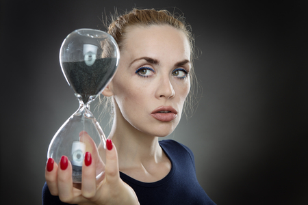 Studio shot of a pretty business model holding a large sandtimer out infront of her.  Black sand is trickling through the hour glass.  Watching the sand of time run out.の写真素材