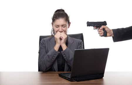 business woman sitting at her desk with a gun to her headの写真素材