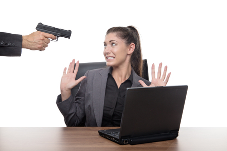 business woman sitting at her desk with a gun to her headの写真素材