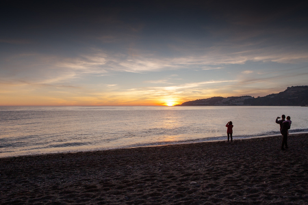 landscape of the sea and beach with people in silhouette taking pitcures on their phone of the beautiful sunsetの写真素材