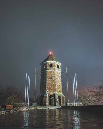 Watchtower pictured on a rainy night. Red light on top of tower. Reflections in the water on the stone walkway. Cloudy sky in background.の写真素材