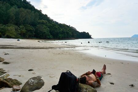 Langkawi, Malaysia - October 12, 2019. Guy lay on Pasir Tengkorak Beach and look on his phoneのeditorial素材