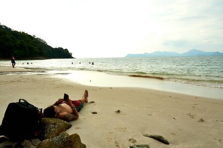 Langkawi, Malaysia - October 12, 2019. Guy lay on Pasir Tengkorak Beach and look on his phoneのeditorial素材