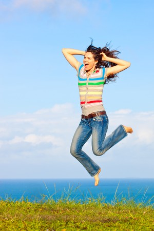 A beautiful young woman jumping up in the air laughing outdoors by a beautiful oceanの写真素材