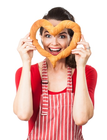A beautiful happy woman wearing an apron is smiling and peeking through a love heart made out of bread  Isolated on white の写真素材