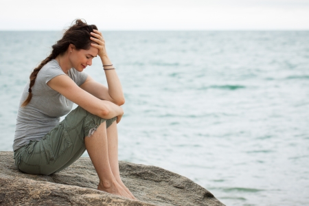 Upset and depressed woman sitting by the ocean crying with her head in her hand の写真素材