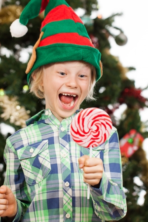 A happy laughing boy dressed as Santas helper or an elf is holding and looking at a big red lollipop. Isolated on white.の写真素材