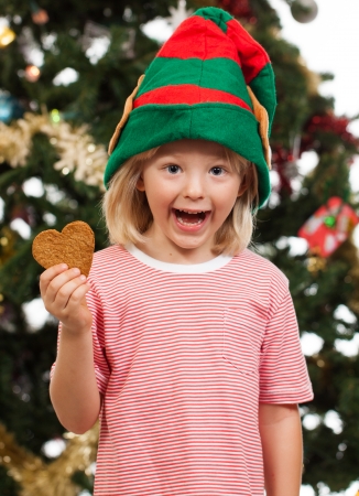 A cute excited boy dressed as Santa's helper is holding gingerbread cookie in front of a Christmas tree.の写真素材