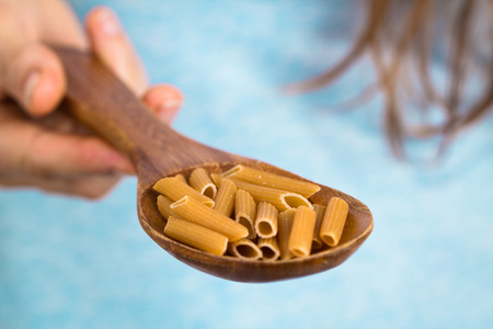 A hand holding a wooden spoon with wholemeal penne pastaの写真素材