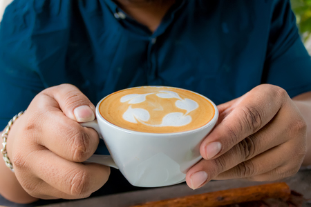 A man holding a glass of coffee latte in hand on a wooden table.の写真素材