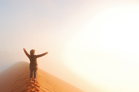 Person standing on top of dune in desert and looking at rising sun in mist with hands up, travel in Africa, Namibiaの写真素材
