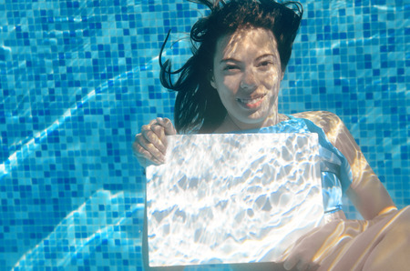 Beautiful young girl holding white blank board in swimming pool under water, fitness and fun on family vacationの写真素材