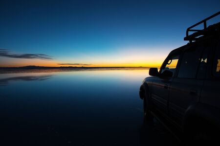 Sunrise on Salar de Uyuni in Bolivia covered with water, car in salt flat desert and sky reflectionsの写真素材