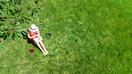 Young woman reading book in park, student girl relaxing outdoors sitting on grass with book and headphones, aerial drone view from aboveの写真素材
