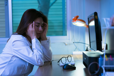 Portrait of young stressed woman sitting at home office desk. Touching head with tired facial expression, at nightの写真素材