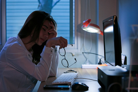Portrait of young stressed woman sitting at home office desk. Touching head with tired facial expression, at nightの写真素材