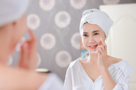 Young asia woman applying foundation or moisturizer on her face in front of mirror.の写真素材